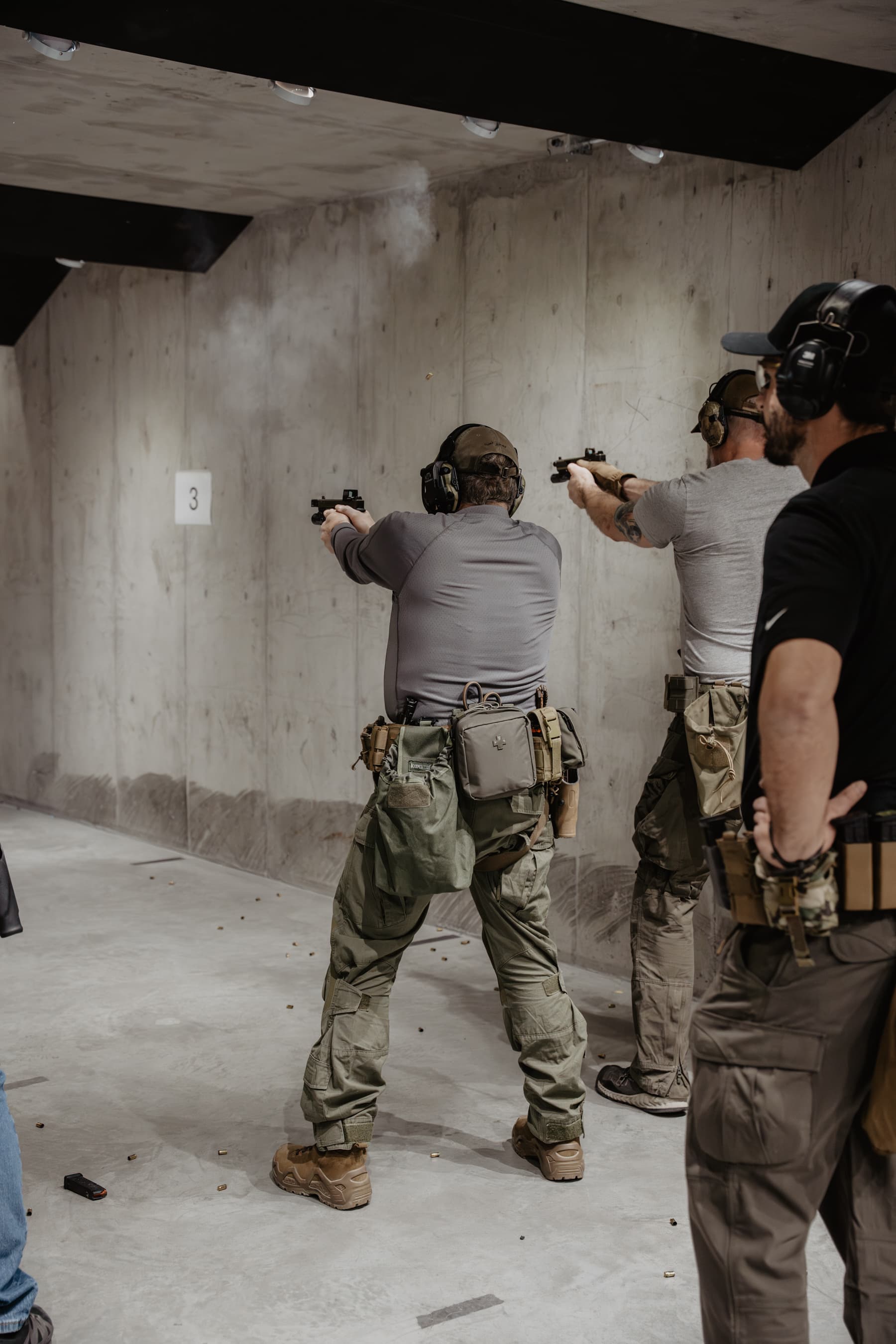 Two students mid-fire on the range, smoke from the muzzle, instructor observing